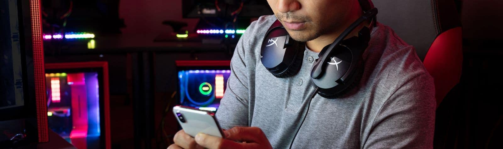 A person wearing a gray hoodie and HyperX gaming headphones around their neck looking at a smartphone in a dimly lit room with RGB gaming setup lighting visible in the background